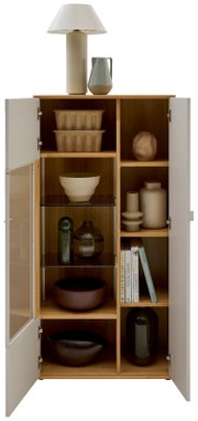 Frontansicht eines offenen, hohen Wohnzimmerschranks aus hellem Holz mit weißen Türen und Glaseinsatz, gefüllt mit Schalen, Vasen und Büchern; oben stehen eine Tischlampe und ein Krug