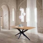 Rechteckiger Esstisch mit heller Holzplatte und schwarzem X-förmigem Metallgestell in einem hellen Raum mit Steinbögen und Hängelampen, Perspektive: leicht seitliche Ansicht auf Tischhöhe