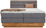 Frontale Ansicht eines Boxspringbetts mit grauem Stoffbezug, hölzernem Sockel, beiger Decke und Kissen.