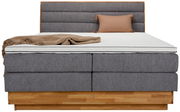 Frontale Ansicht eines Boxspringbetts mit grauem Stoffbezug, hölzernem Sockel, beiger Decke und Kissen.