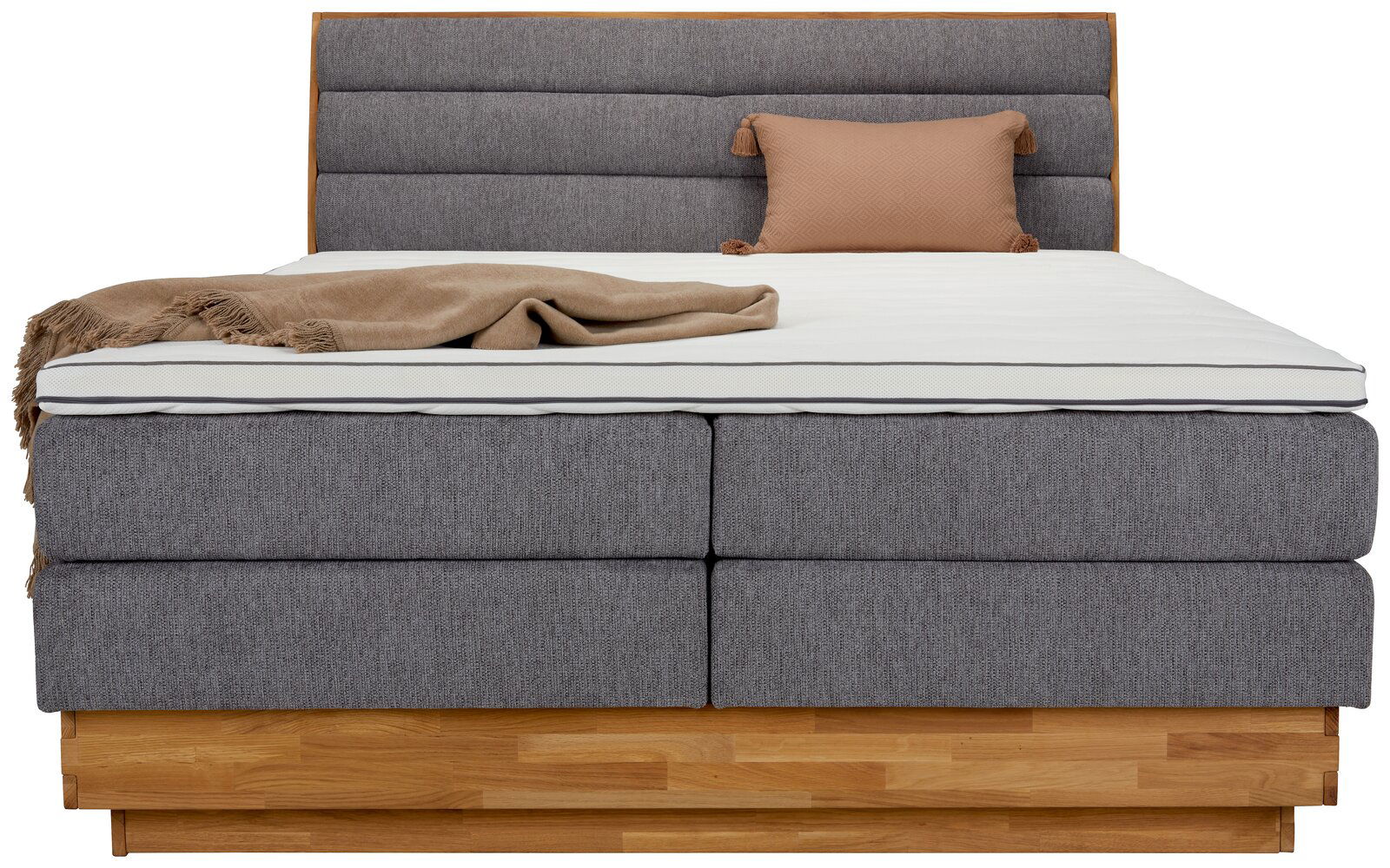 Frontale Ansicht eines Boxspringbetts mit grauem Stoffbezug, hölzernem Sockel, beiger Decke und Kissen.