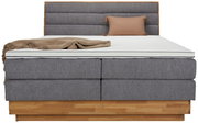 Frontale Ansicht eines Boxspringbetts mit grauem Stoffbezug, hölzernem Sockel, beiger Decke und Kissen.