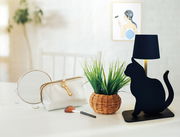 Schwarze Akku-Tischleuchte in Katzenform, seitliche Perspektive, auf einem Tisch neben einem Spiegel, einer Kosmetiktasche und einem Pinsel.