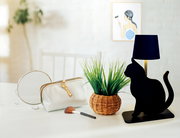 Schwarze Akku-Tischleuchte in Katzenform, seitliche Perspektive, auf einem Tisch neben einem Spiegel, einer Kosmetiktasche und einem Pinsel.