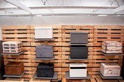 Frontalansicht einer Ausstellungswand aus Holzpaletten mit mehreren quadratischen Sitzhockern und Aufbewahrungsboxen in verschiedenen Farben und Stoffen auf grauen Wandregalen