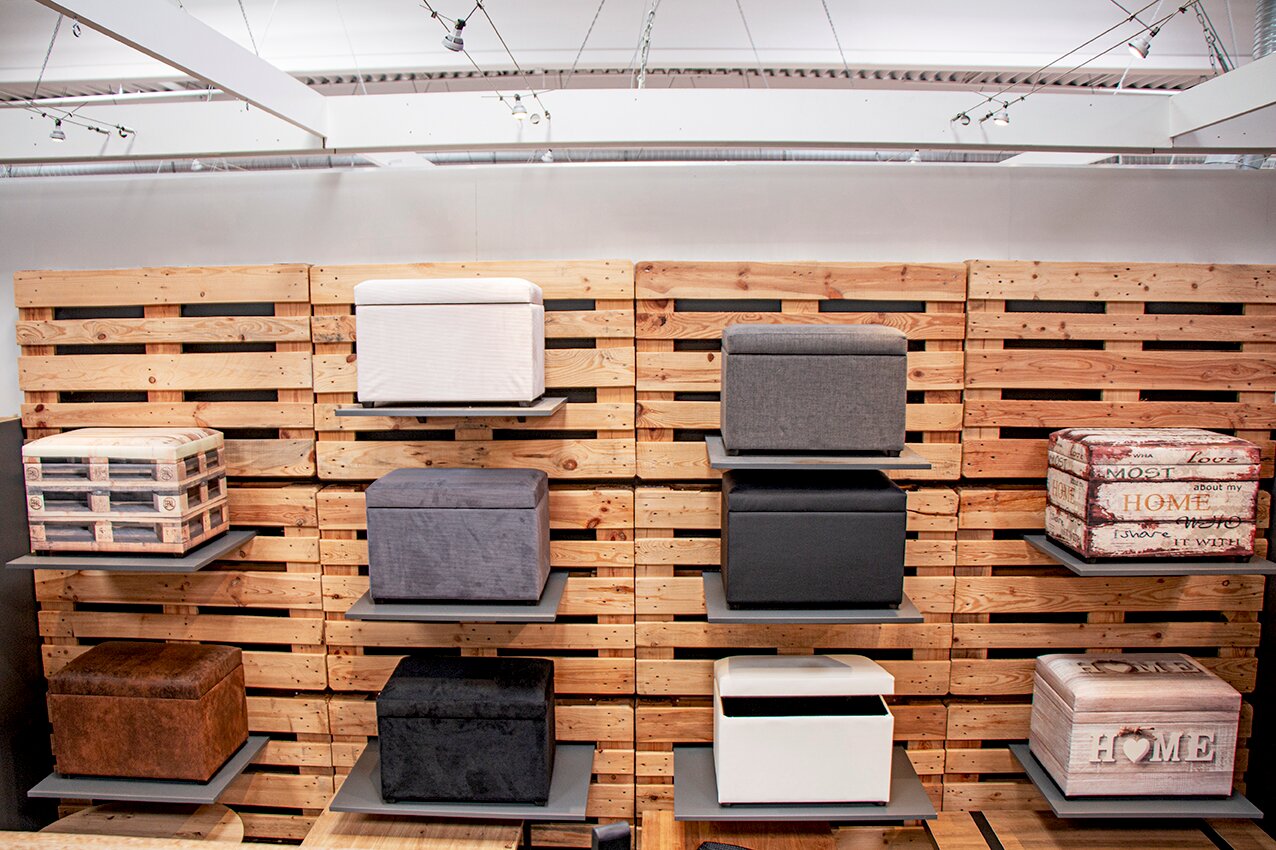 Frontalansicht einer Ausstellungswand aus Holzpaletten mit mehreren quadratischen Sitzhockern und Aufbewahrungsboxen in verschiedenen Farben und Stoffen auf grauen Wandregalen