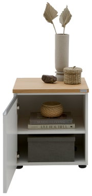 Weißer Nachttisch/Beistellschrank mit geöffneter Tür, Holzplatte und Metallgriff; oben Vase und Deko, innen Regalfach mit Büchern und Box – frontale Ansicht leicht von oben.