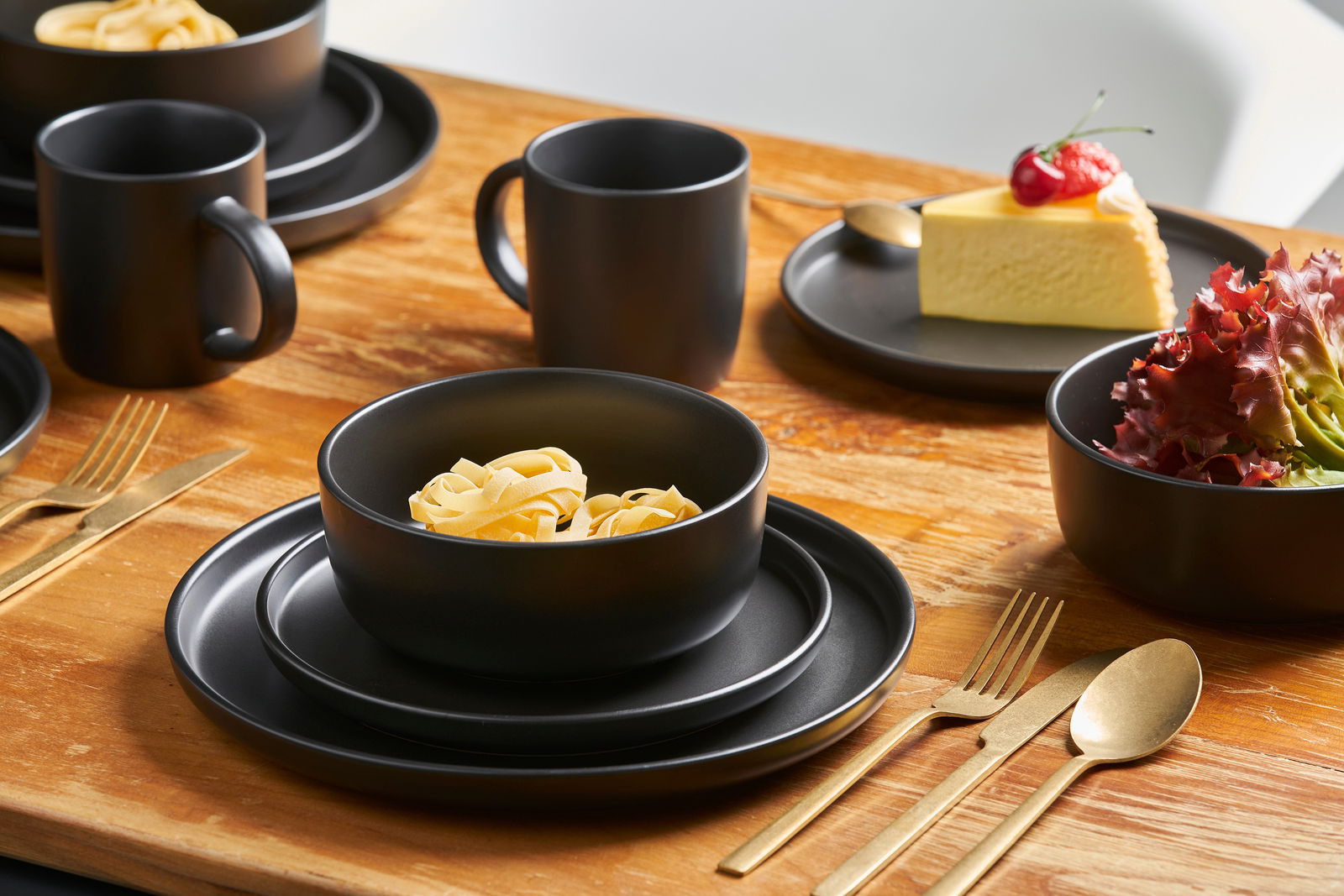 Schwarzes Kombiservice Nero 16-tlg. auf einem Holztisch, bestehend aus Tellern, Schalen und Tassen, mit goldfarbenem Besteck, aus der Perspektive von schräg oben.