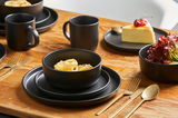 Schwarzes Kombiservice Nero 16-tlg. auf einem Holztisch, bestehend aus Tellern, Schalen und Tassen, mit goldfarbenem Besteck, aus der Perspektive von schräg oben.