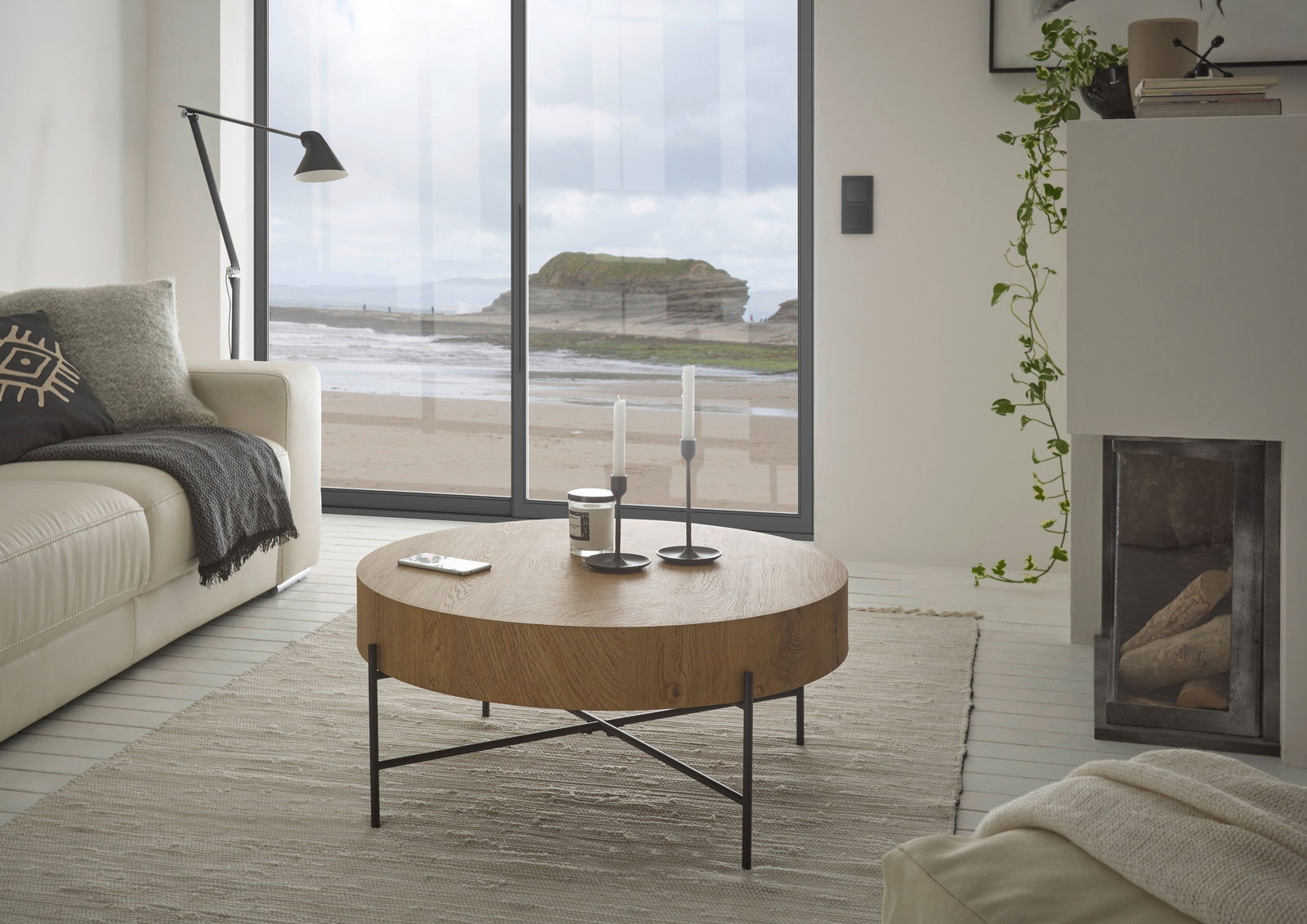 Runder Couchtisch aus Holz mit schwarzem Metallgestell in einem modernen Wohnzimmer mit Blick auf das Meer durch große Fenster.