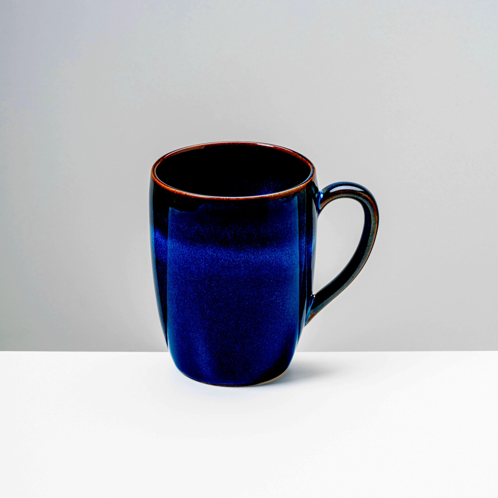 Dunkelblaue, glänzende Keramik-Kaffeetasse mit Henkel vor hellem Hintergrund, frontal aus leicht erhöhter Perspektive aufgenommen