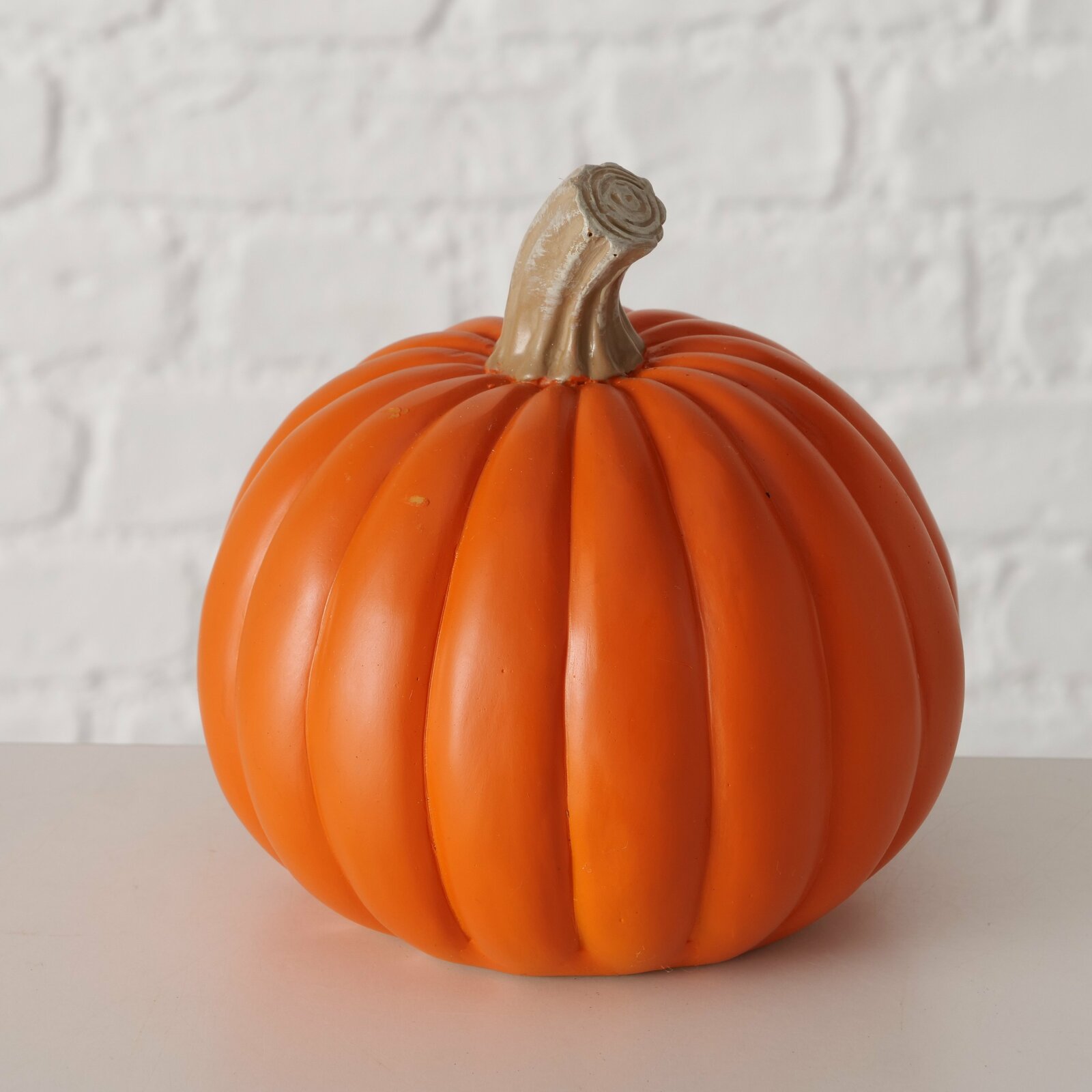 Dekorativer Kürbis aus Kunststoff, frontal fotografiert vor einer weißen Ziegelwand.