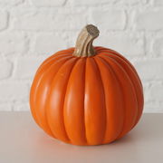 Dekorativer Kürbis aus Kunststoff, frontal fotografiert vor einer weißen Ziegelwand.