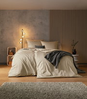Gemütliches Schlafzimmer mit einem großen Bett in beiger Bettwäsche, dekoriert mit grauen Kissen und einer Decke. Im Hintergrund eine moderne Stehlampe und eine stilvolle Wandverkleidung. Perspektive von vorne.