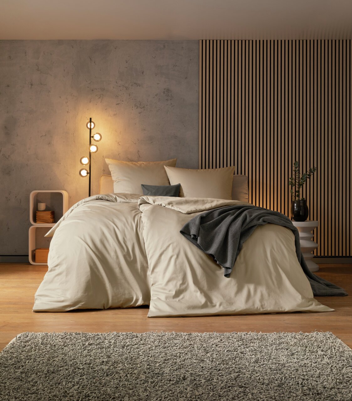 Gemütliches Schlafzimmer mit einem großen Bett in beiger Bettwäsche, dekoriert mit grauen Kissen und einer Decke. Im Hintergrund eine moderne Stehlampe und eine stilvolle Wandverkleidung. Perspektive von vorne.