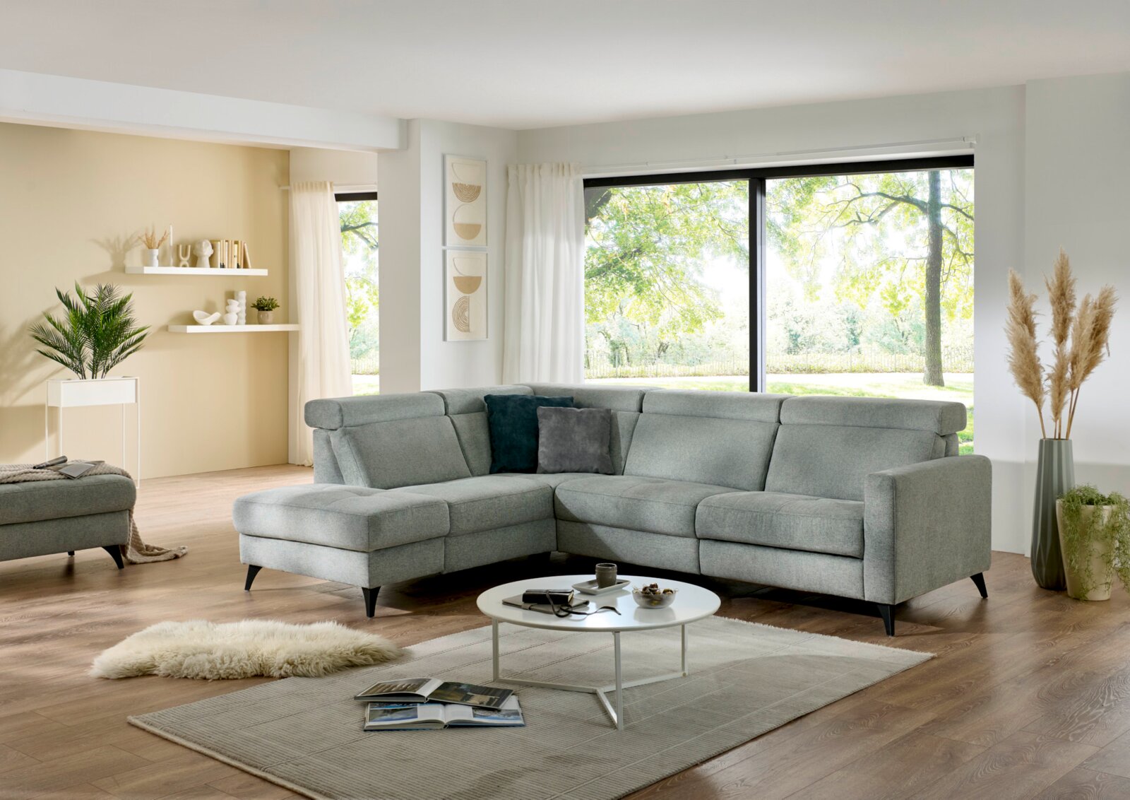 Modernes graues L-förmiges Ecksofa mit Ottomane links und schwarzen Füßen in hellem Wohnzimmer, Frontansicht leicht schräg von vorn.
