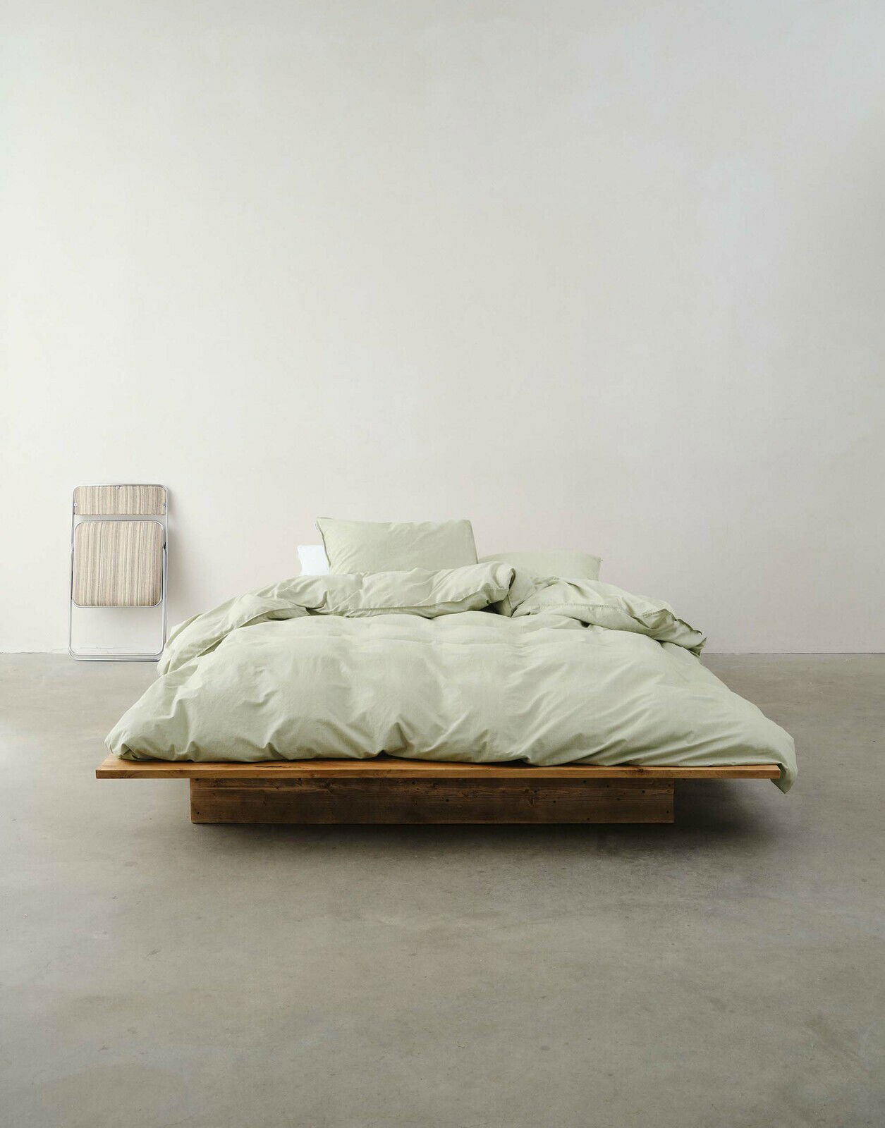 Minimalistisches Schlafzimmer mit einem niedrigen Holzbett, bezogen mit hellgrüner Bettwäsche, aus der Vorderansicht fotografiert.