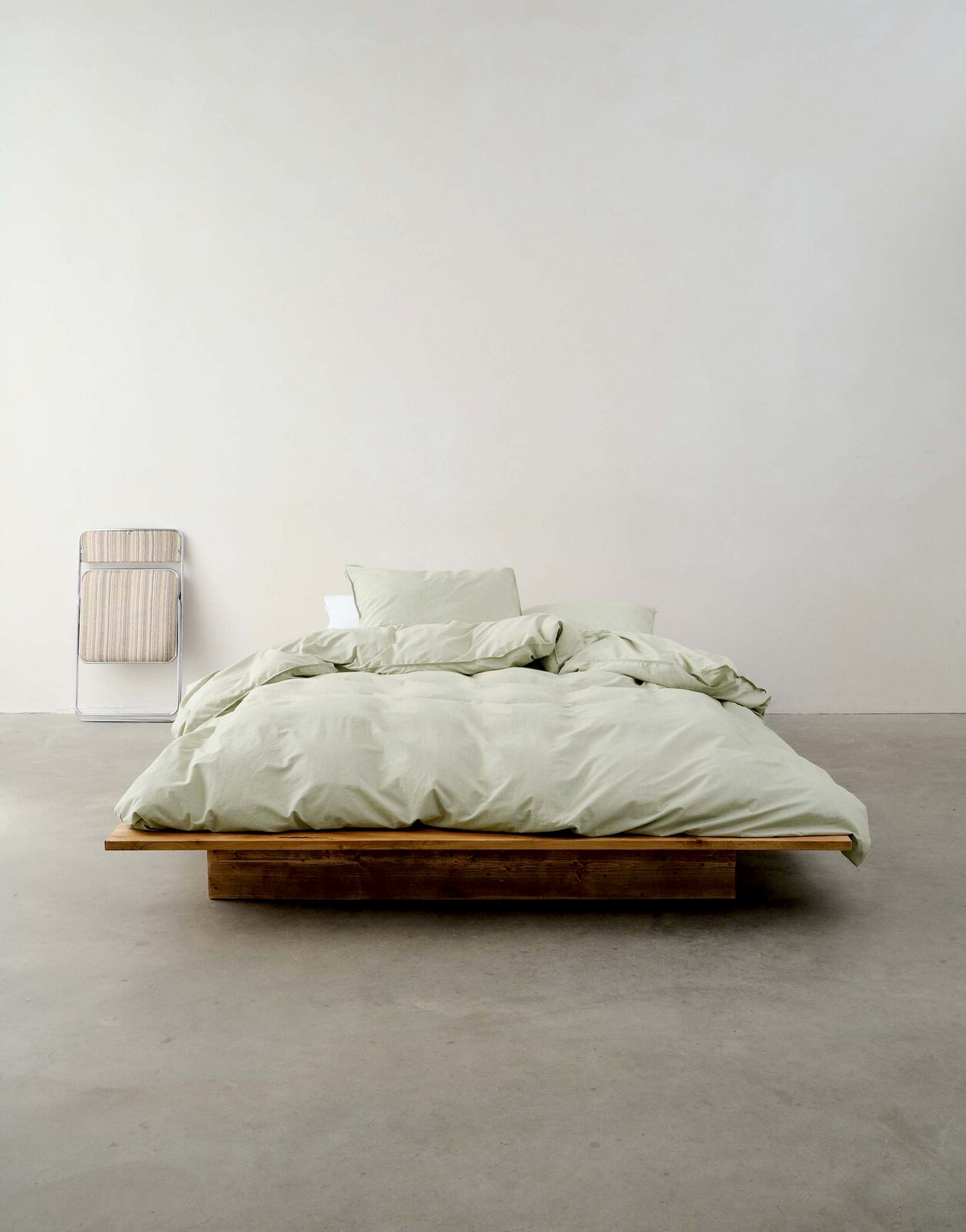 Minimalistisches Schlafzimmer mit einem niedrigen Holzbett, bezogen mit hellgrüner Bettwäsche, aus der Vorderansicht fotografiert.