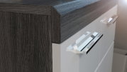 Nahaufnahme aus schräger Seitenperspektive: moderner Schrank/Kommode mit dunkler Holzoptik am Korpus, weißer Hochglanzfront und verchromten Griffen