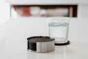 Runder Untersetzer-Set aus Edelstahl mit schwarzen Einlagen auf weißem Tisch, daneben ein Glas Wasser auf einem Untersetzer, fotografiert in leichter Seitenansicht auf Tischhöhe