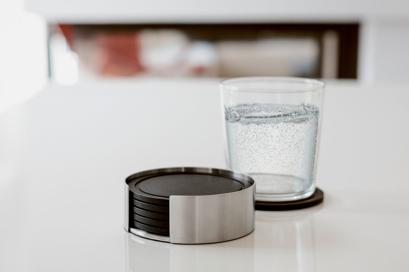 Runder Untersetzer-Set aus Edelstahl mit schwarzen Einlagen auf weißem Tisch, daneben ein Glas Wasser auf einem Untersetzer, fotografiert in leichter Seitenansicht auf Tischhöhe