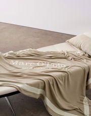 Beige Plaid 150x200cm mit dem Aufdruck 'Marc O'Polo EST. IN STOCKHOLM', drapiert auf einem Sofa, seitliche Perspektive.