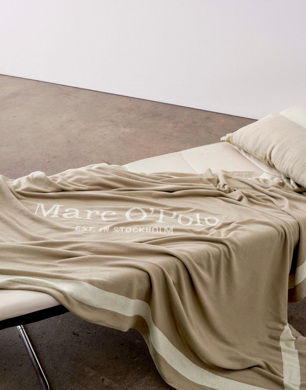 Beige Plaid 150x200cm mit dem Aufdruck 'Marc O'Polo EST. IN STOCKHOLM', drapiert auf einem Sofa, seitliche Perspektive.