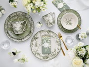 Draufsicht auf ein elegantes Tischgedeck mit grün-weißem Porzellan im floralen Vogel-Design (Teller, Schale und Tasse), goldfarbenem Besteck, Serviette und weißen Blumen auf hellem Hintergrund.