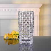Transparente Vase mit quadratischem Muster, frontal fotografiert, auf einem Tisch mit gelben Blumen im Hintergrund.