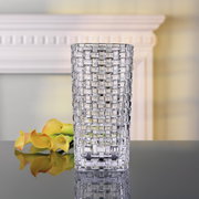 Transparente Vase mit quadratischem Muster, frontal fotografiert, auf einem Tisch mit gelben Blumen im Hintergrund.