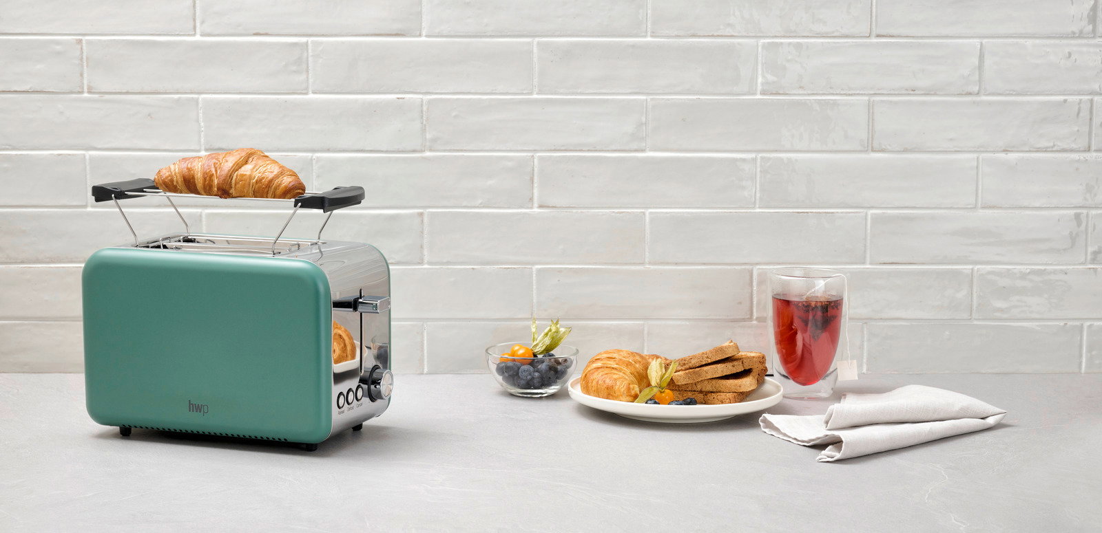 Grüner Toaster mit Croissant auf dem Brötchenaufsatz, seitliche Perspektive, neben einem Teller mit Toast und Beeren.
