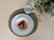 Draufsicht auf ein weißes Geschirr-Set mit Teller und Schale auf grünem Platzset, daneben eine weiße Tasse; in der Schale Müsli mit roten Johannisbeeren, rechts kleine Kekse und oben rechts Blumen.