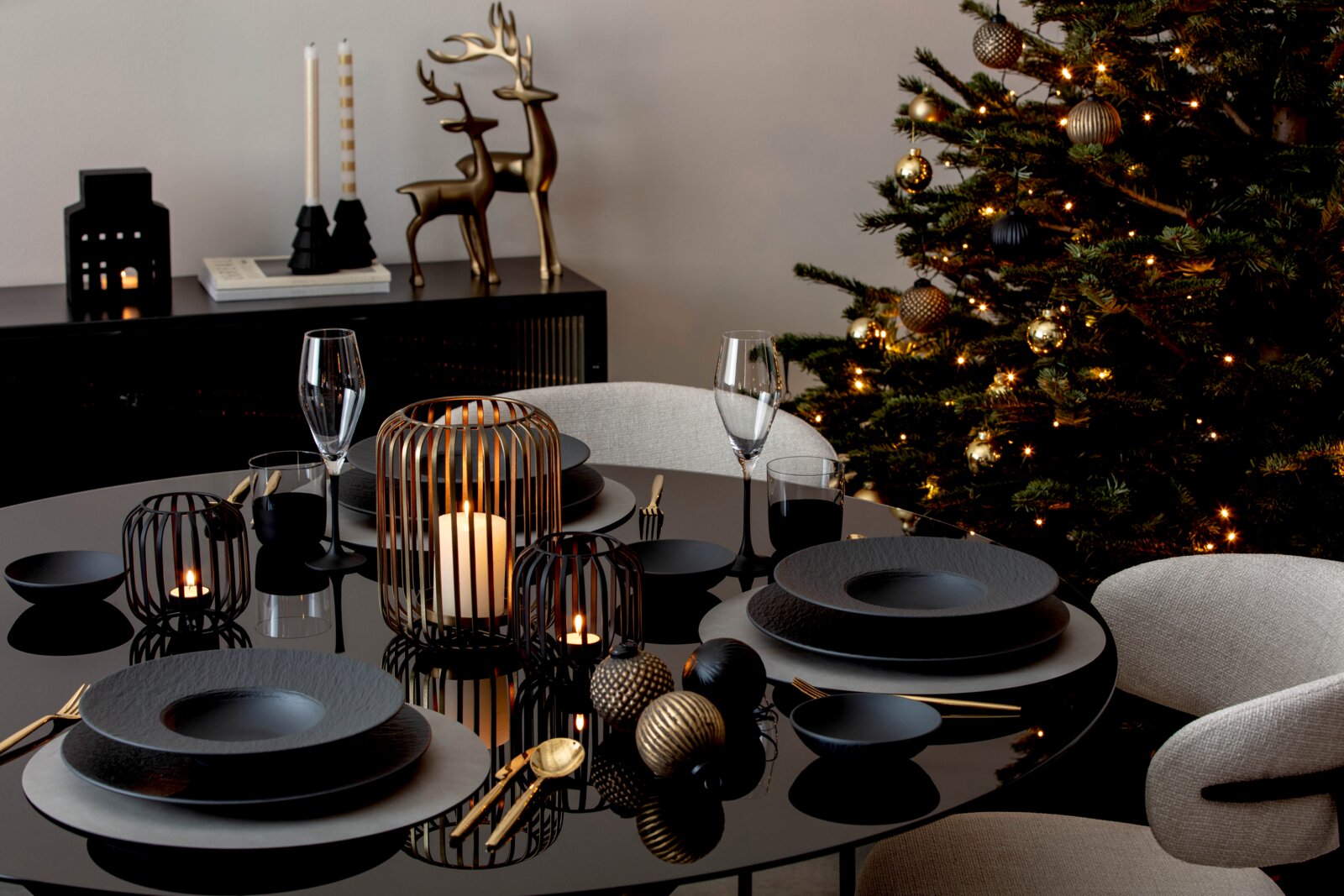 Villeroy & Boch Weihnachtskugeln 3er-Set Festlich gedeckter Esstisch in Schwarz und Gold mit edlem Geschirr, Kerzenlaternen und Weihnachtskugeln vor einem beleuchteten Weihnachtsbaum, aus leicht erhöhter Frontalperspektive