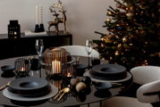 Festlich gedeckter Esstisch in Schwarz und Gold mit edlem Geschirr, Kerzenlaternen und Weihnachtskugeln vor einem beleuchteten Weihnachtsbaum, aus leicht erhöhter Frontalperspektive