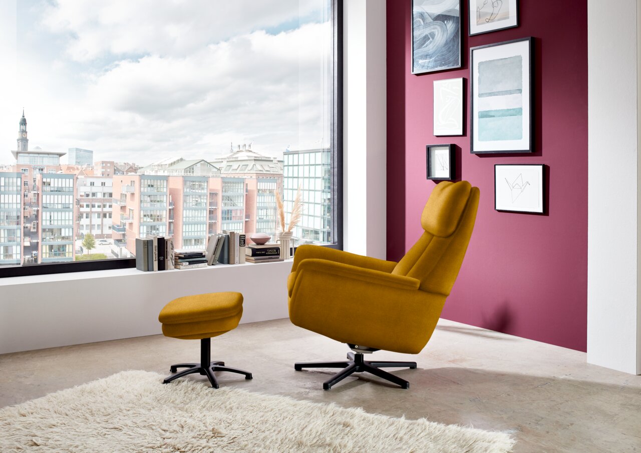 Gele fauteuil met bijpassend voetbankje in een moderne woonkamer, gezien vanuit de zijkant, met uitzicht op grote ramen en de stad.