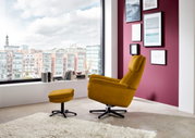 Gele fauteuil met bijpassend voetbankje in een moderne woonkamer, gezien vanuit de zijkant, met uitzicht op grote ramen en de stad.