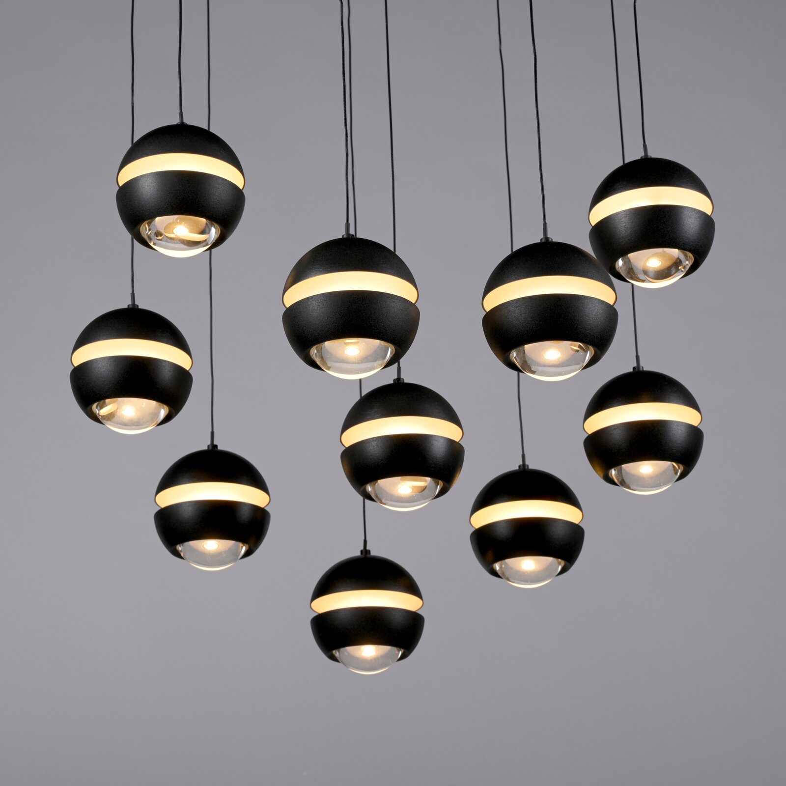Verschillende moderne, bolvormige zwarte hanglampen met een rondomlopende warmwitte lichtband en een glazen lens hangen aan kabels tegen een grijze achtergrond, gezien vanuit een lichte onderhoek (onderzicht).