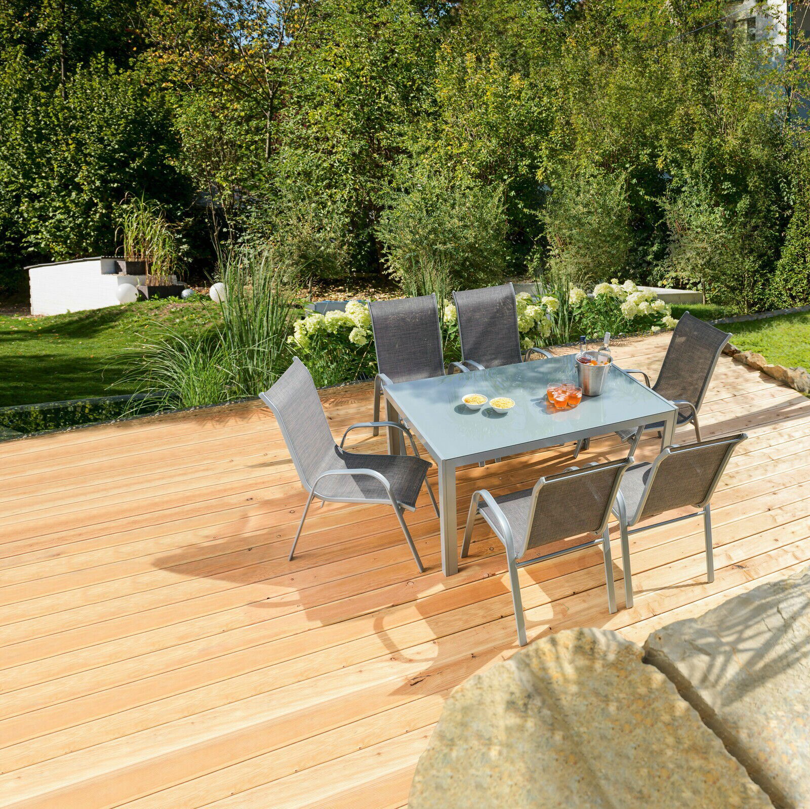 Moderner Gartentisch mit Glasplatte und sechs Stühlen auf einer Holzterrasse, umgeben von grüner Gartenlandschaft, aus der Vogelperspektive fotografiert.