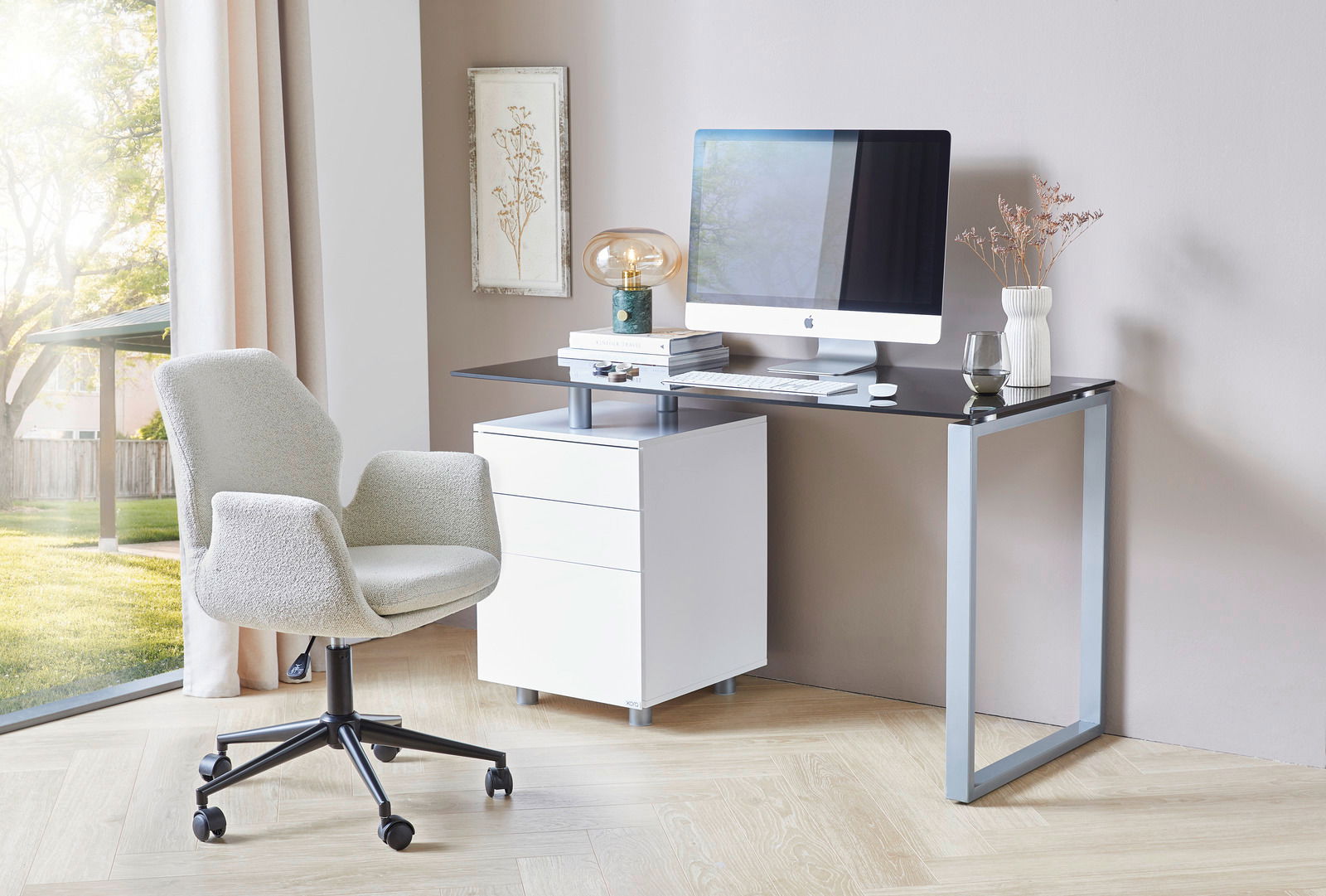 Moderner Schreibtisch mit Glasplatte und Metallrahmen, ausgestattet mit einem weißen Rollcontainer und einem Computer, seitliche Perspektive.