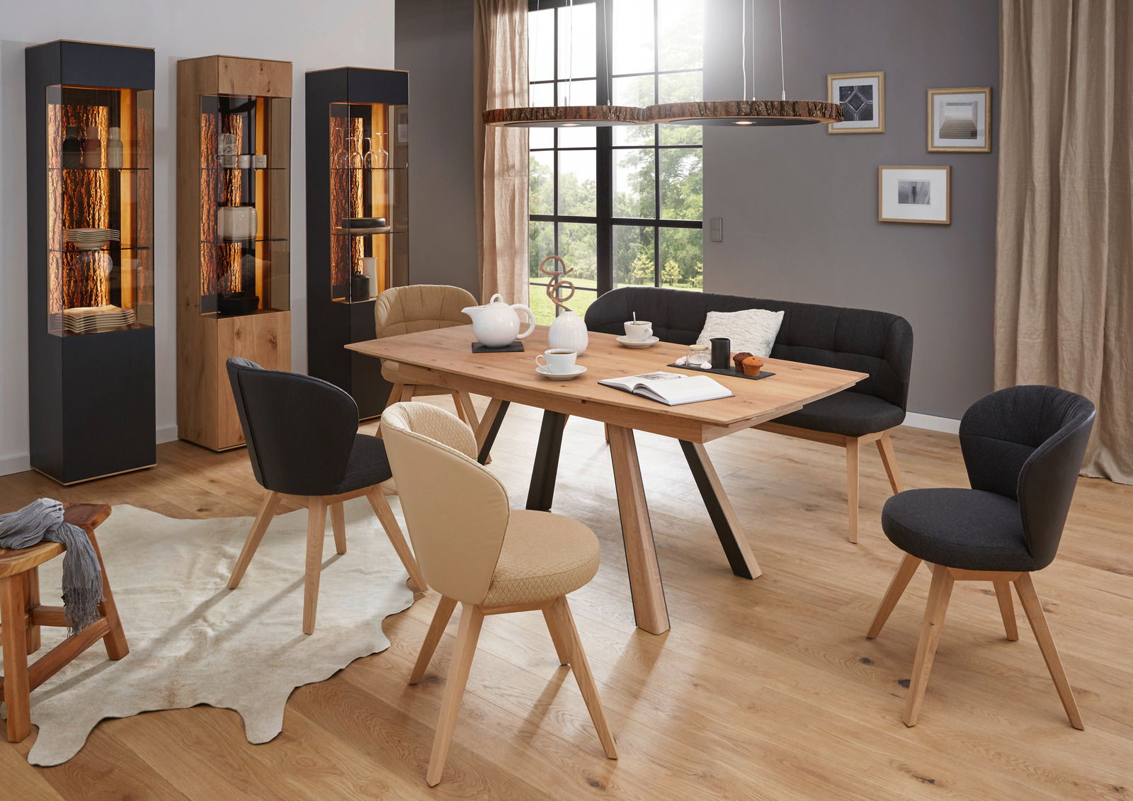 Moderne Esszimmer-Szene mit Valnatura Vitrine LAVIA links im Bild, aus Holz und Glas, beleuchtet, neben einem Esstisch mit Stühlen und einer Bank.