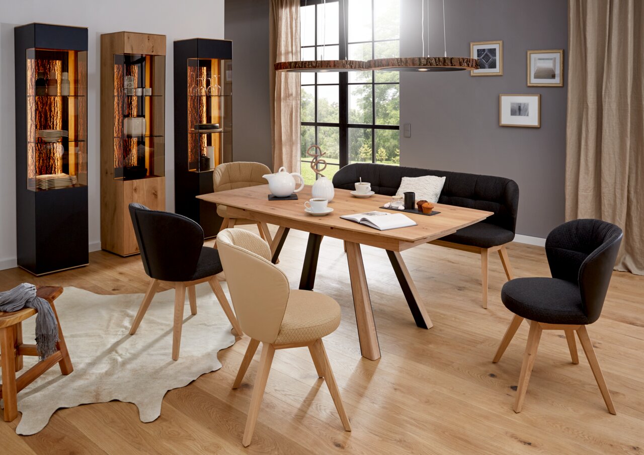 Moderne Esszimmer-Szene mit Valnatura Vitrine LAVIA links im Bild, aus Holz und Glas, beleuchtet, neben einem Esstisch mit Stühlen und einer Bank.