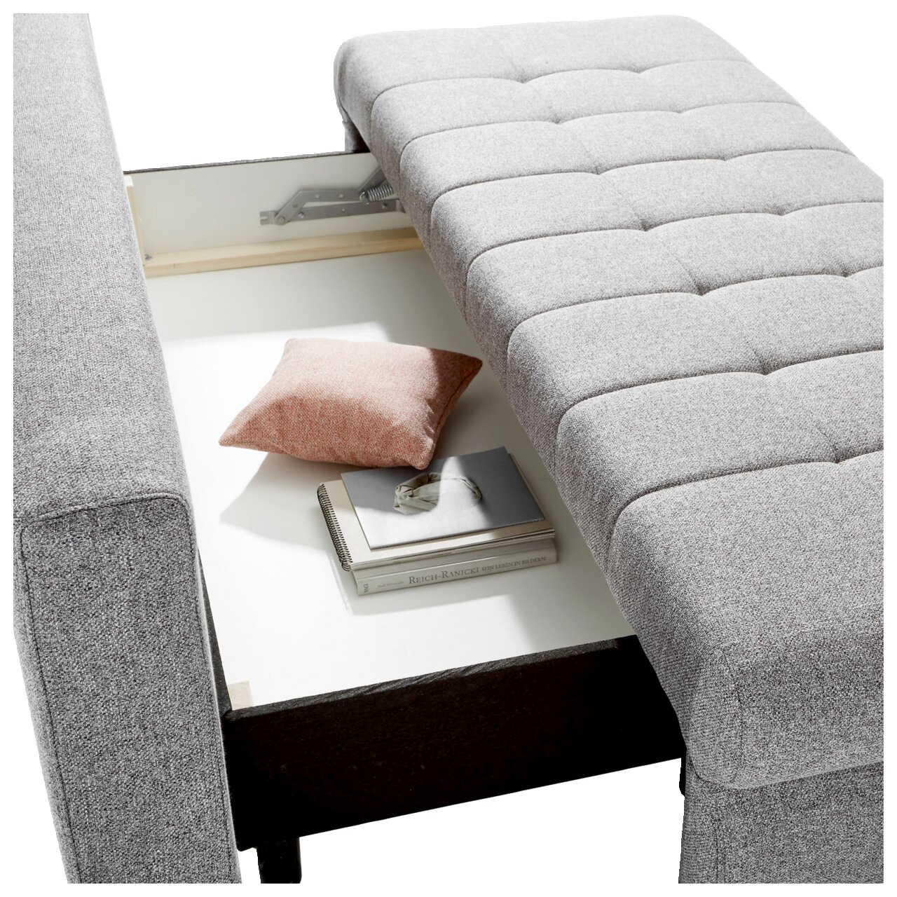 Graues Funktionssofa mit geöffneter Aufbewahrungsbox, von oben fotografiert. Ein rosa Kissen und Bücher sind im Stauraum sichtbar.