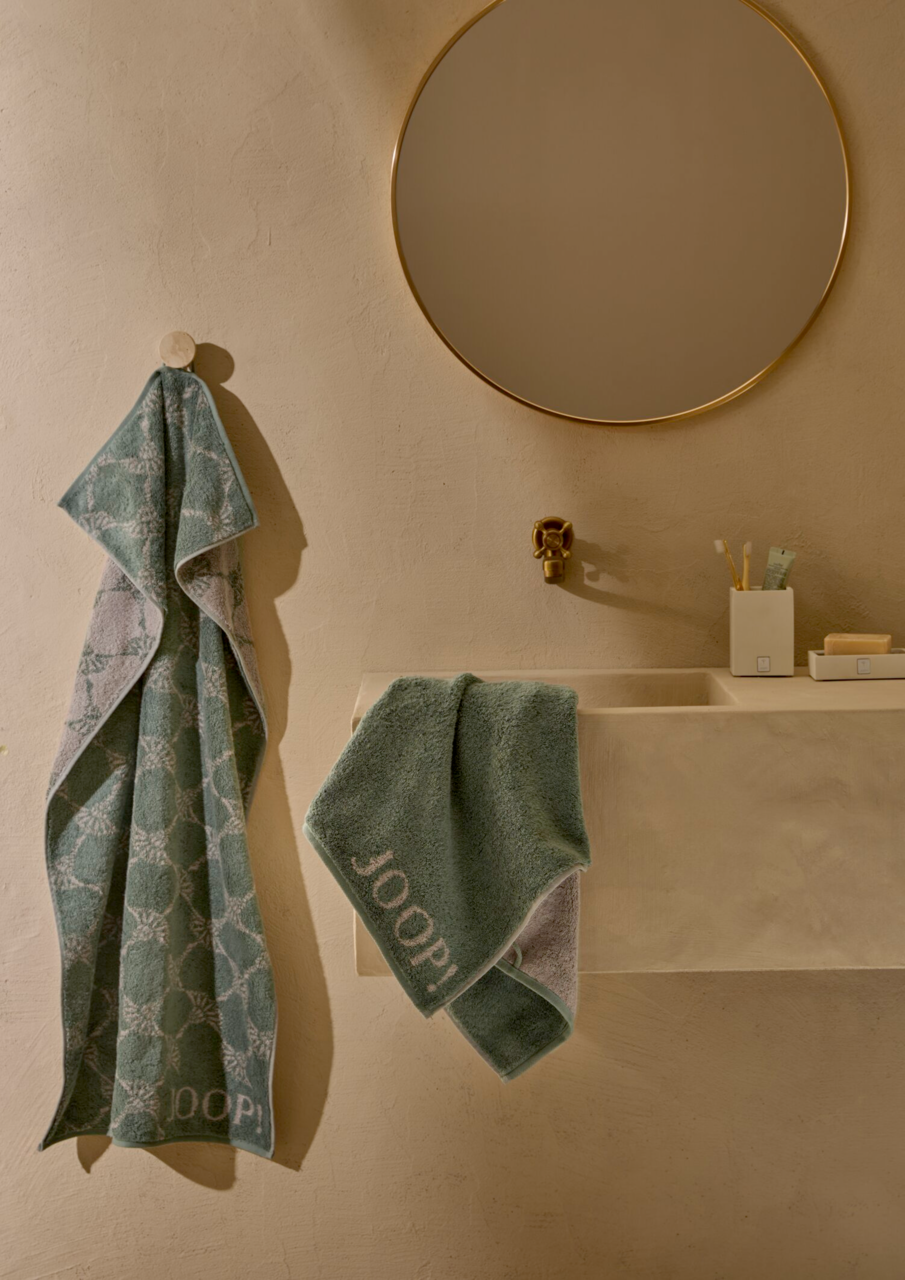 Zwei grüne Handtücher mit Muster, eines hängt an einem Haken, das andere liegt auf einem Waschbecken. Perspektive: Frontalansicht.
