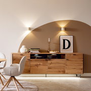 Modernes Wohnzimmer-Setting mit breitem Sideboard aus hellem Holz, Deko und zwei Wandleuchten vor zweifarbig gestrichener Wand; frontale Perspektive auf das Möbelstück.
