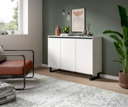 Modernes weißes Sideboard mit drei Türen, schwarzem Sockel und Dekorationen, in einem stilvoll eingerichteten Wohnzimmer, seitliche Perspektive.