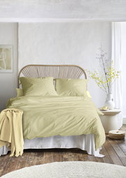 Gemütliches Schlafzimmer mit einem Bett, das mit hellgelber Bettwäsche bezogen ist, aus der Vorderansicht fotografiert. Im Hintergrund ein Rattan-Kopfteil und ein Nachttisch mit Vase und Blumen.