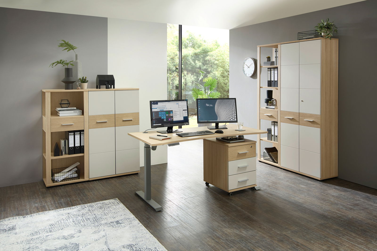 Moderner Büroraum mit einem höhenverstellbaren Schreibtisch, ausgestattet mit zwei Monitoren, Tastatur und Maus. Im Hintergrund sind Aktenschränke und Regale zu sehen. Die Perspektive zeigt den Raum von vorne.