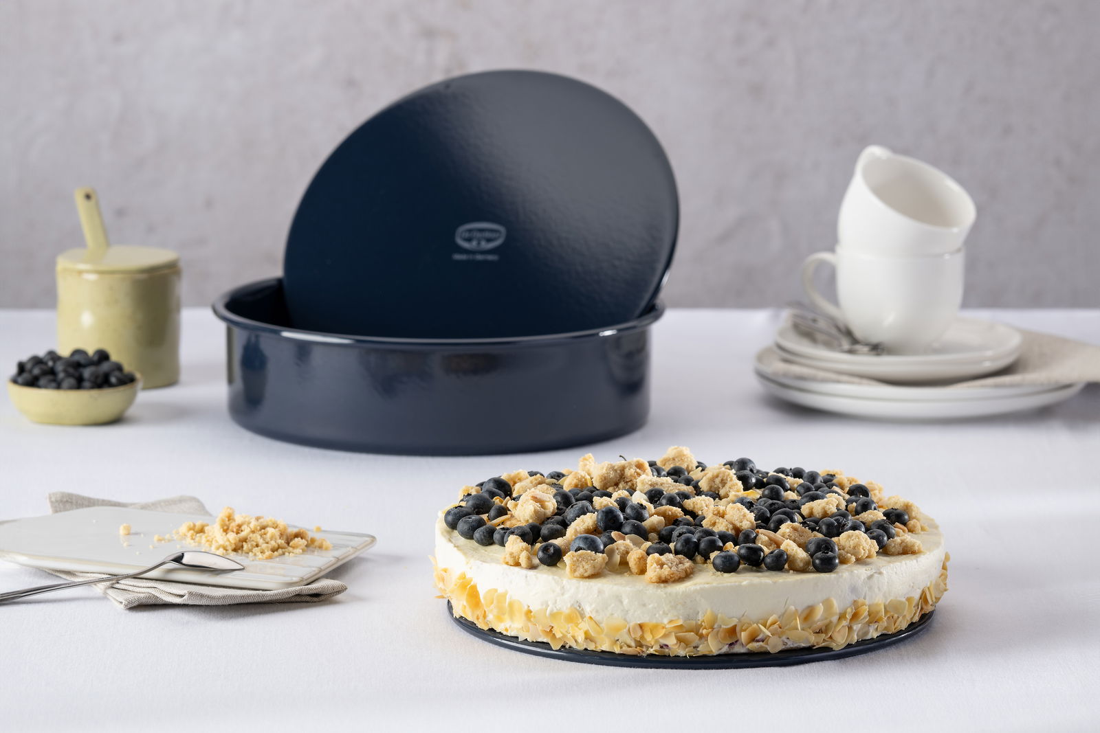 Dr. Oetker Garantieform BACKLIEBE Runde Backform mit abgenommenem Deckel, gefüllt mit einer dekorativen Torte, im Vordergrund auf einem Tisch mit Geschirr und Beeren, seitliche Perspektive.