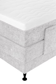 Close-up in schuin bovenaanzicht van een lichtgrijze gestoffeerde boxspring met wit, golvend doorgestikt matras en zwarte poot.