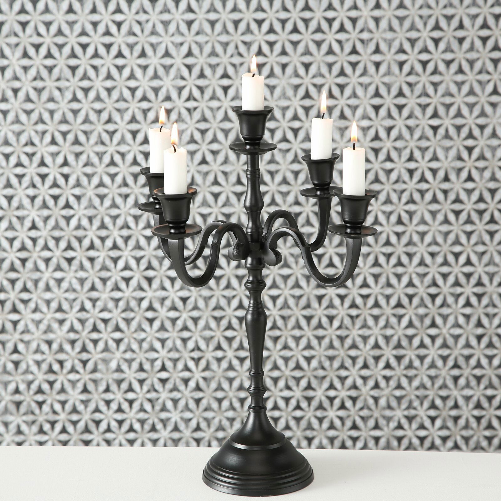 Schwarzer Kerzenleuchter Victoria mit fünf Armen und brennenden Kerzen, frontal vor gemustertem Hintergrund.